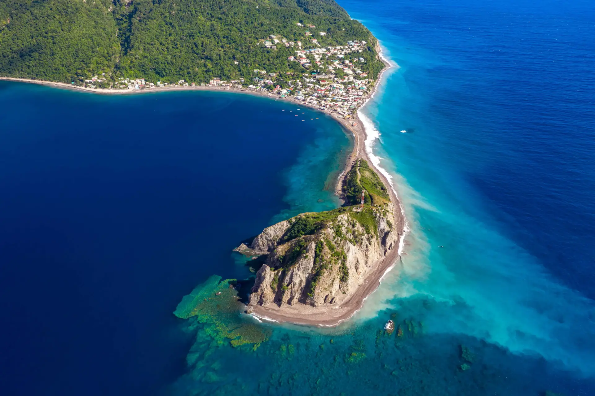 A striking bird's-eye view of a coastal landscape in Dominica. A thin, curved strip of beach and road connects a densely populated coastal village nestled against steep, jungle-covered mountains to a rocky, green headland in the foreground. The water to the left of the land bridge is a calm, dark blue, while the ocean to the right transitions into brilliant shades of light turquoise and aquamarine with visible coral reefs below the surface.