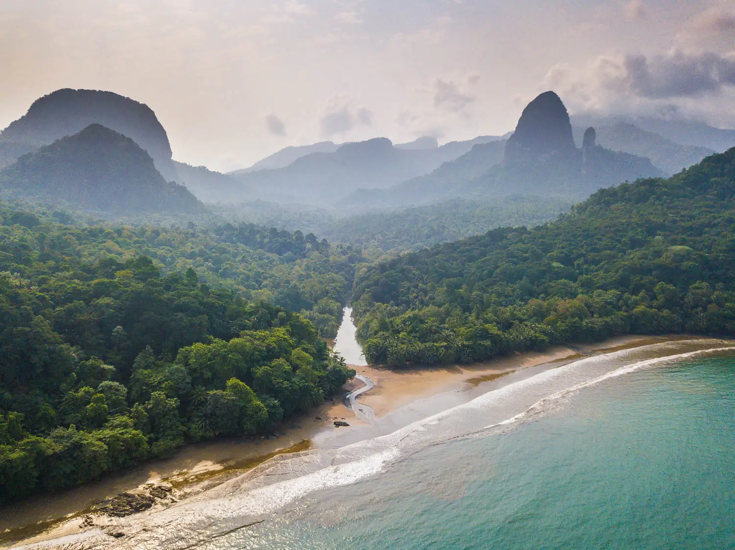 A beautiful high-angle shot looking down a tropical coastline in São Tomé and Príncipe. In the foreground, gentle turquoise waves lap against a curved, sandy beach. A small, winding river breaks through a thick canopy of vibrant green rainforest trees to flow into the sea. The landscape stretches back into a deep, forested valley engulfed in light mist, dominated by striking, steep volcanic rock spires and peaks silhouetted against a hazy, bright sky.