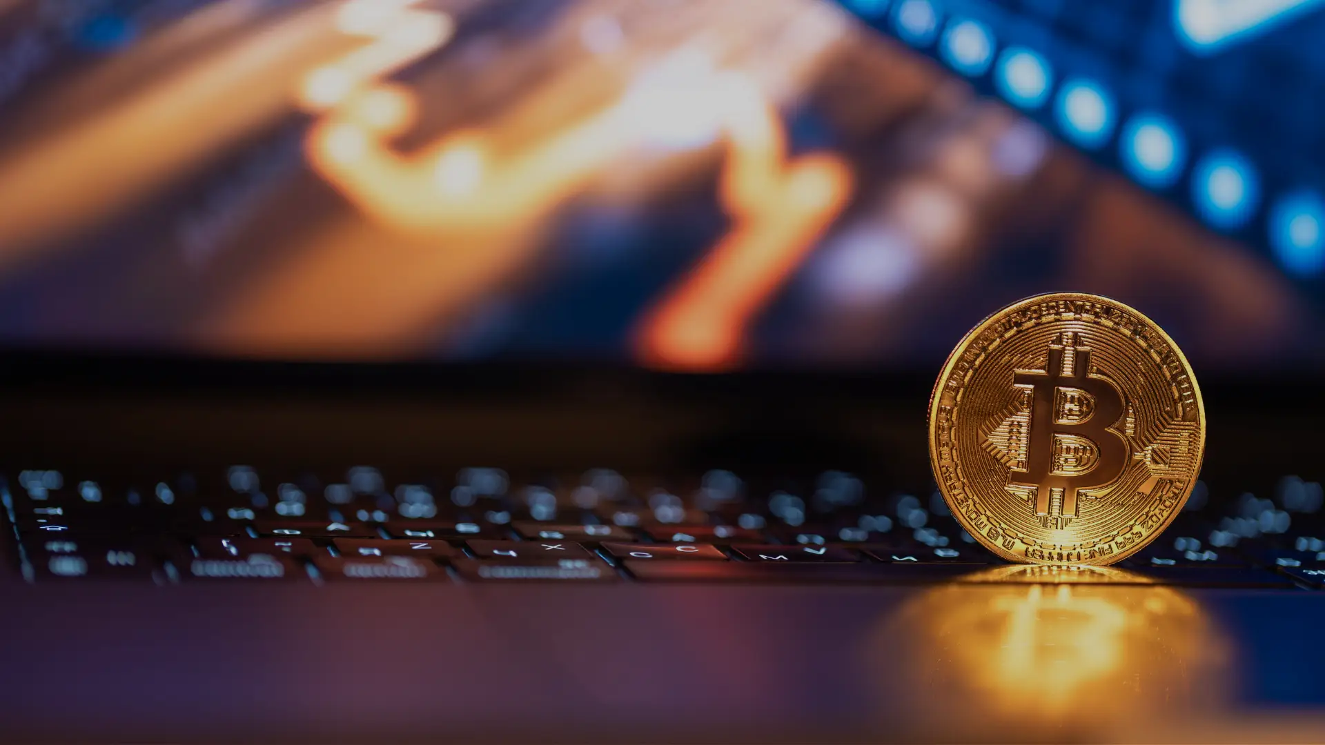 A physical gold Bitcoin coin standing on a laptop keyboard with blurred trading charts displayed on the screen.