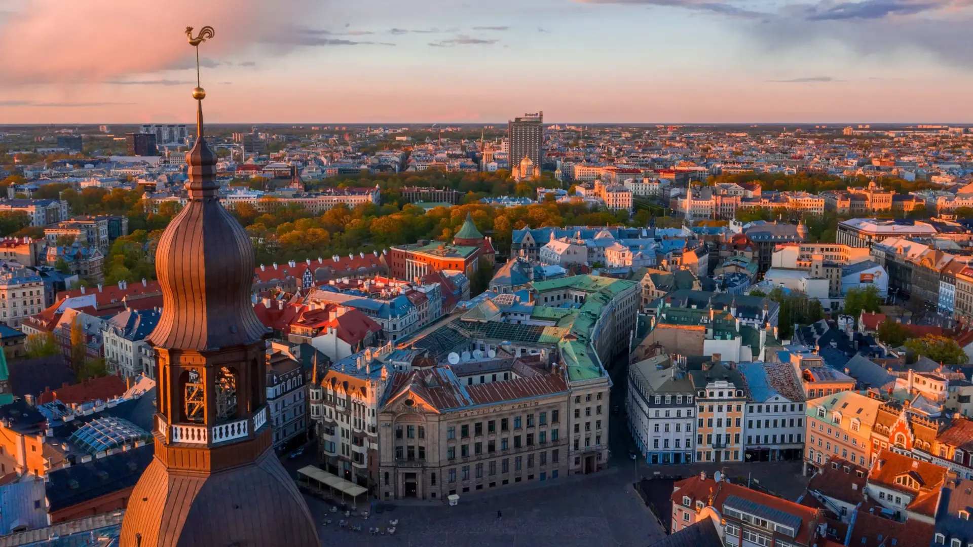 Latvia Golden Visa Update: 2026 Law Changes and Current Routes 1 A beautiful sunset panoramic view of Riga, Latvia. The foreground is dominated on the left by the detailed, tiered wooden architecture of a church spire topped with a distinctive golden rooster. Below, the sprawling historic town square is lined with multi-story European buildings featuring red-orange roofs, teal-accented roofs, and classic facades catching the warm evening light. A dense band of lush green trees cuts through the middle ground, separating the old city from the modern skyline in the distance under a soft orange and pink sky.